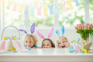 Kids dyeing Easter eggs. Children in bunny ears dye colorful egg for Easter hunt. Home decoration with flowers, basket and rabbit for spring holiday celebration. Little boy and girl decorate home.